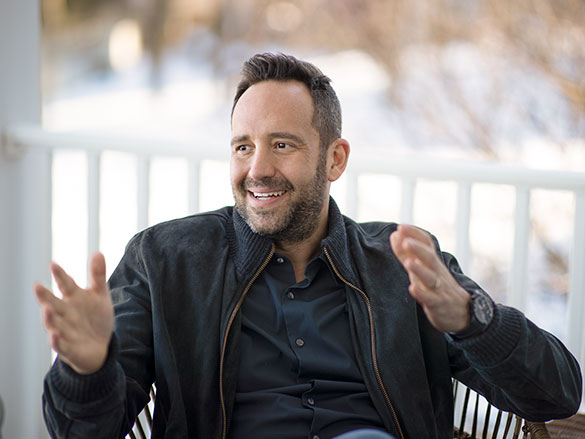 Man wearing a dark jacket sitting down, signaling and smiling