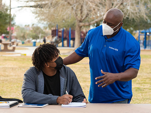 Man standing-up talking to a teenager sitting down on a park table with a pen and notebook 